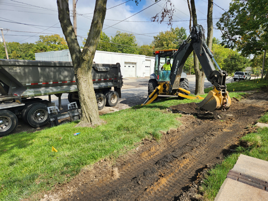 Worker in backhoe digging up dirt where a sidewalk will be installed with a tractor-trailer next to it